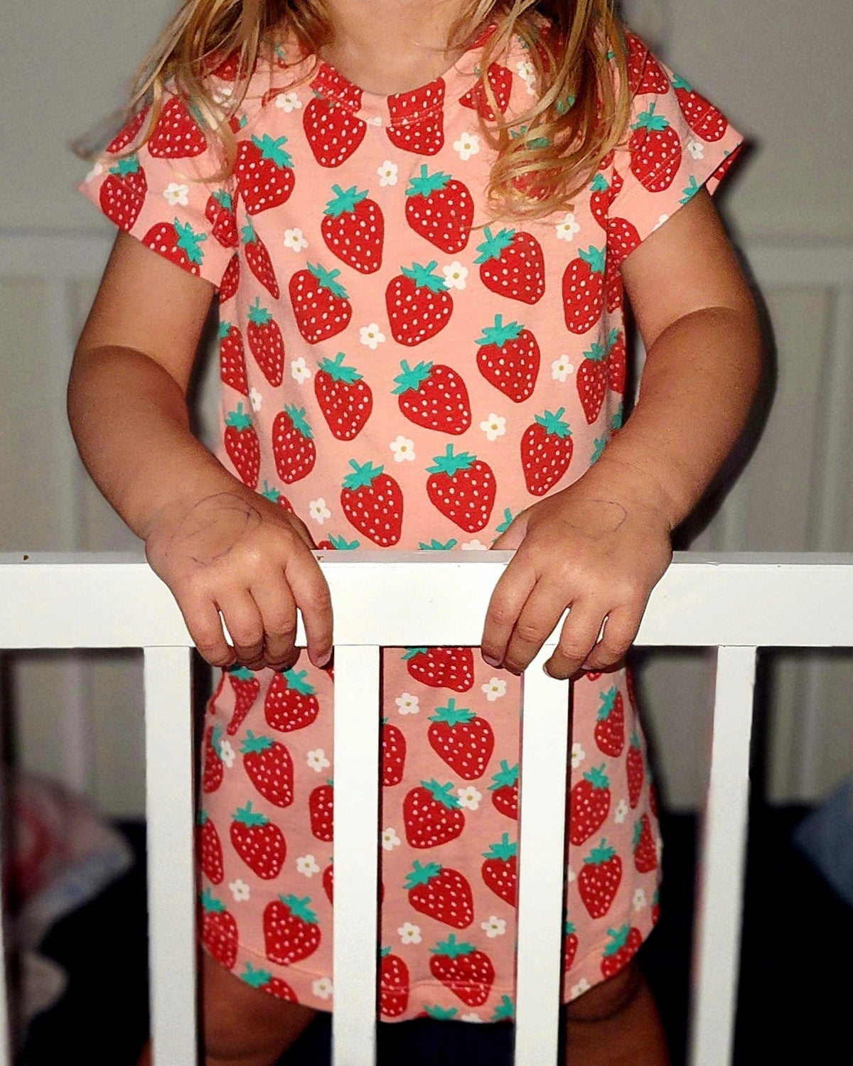 Child wearing a dress with strawberry pattern standing behind a white railing.