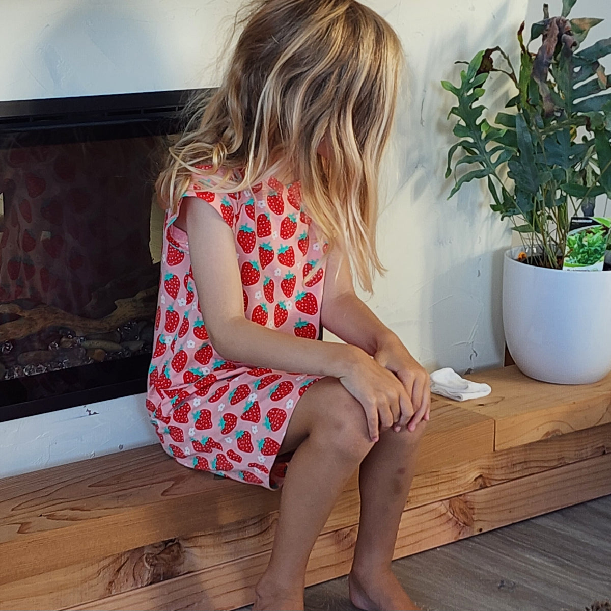 Child wearing a strawberry dress sitting on a wooden bench indoors.