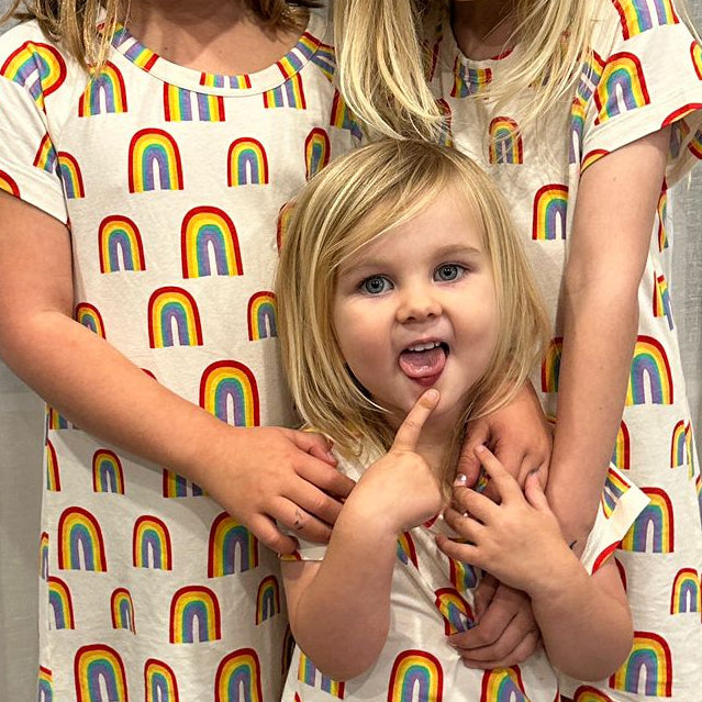 Three children in matching rainbow-patterned dresses standing in front of a white curtain.